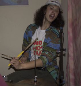 A man in a polaroid baseball cap playing the drums.
